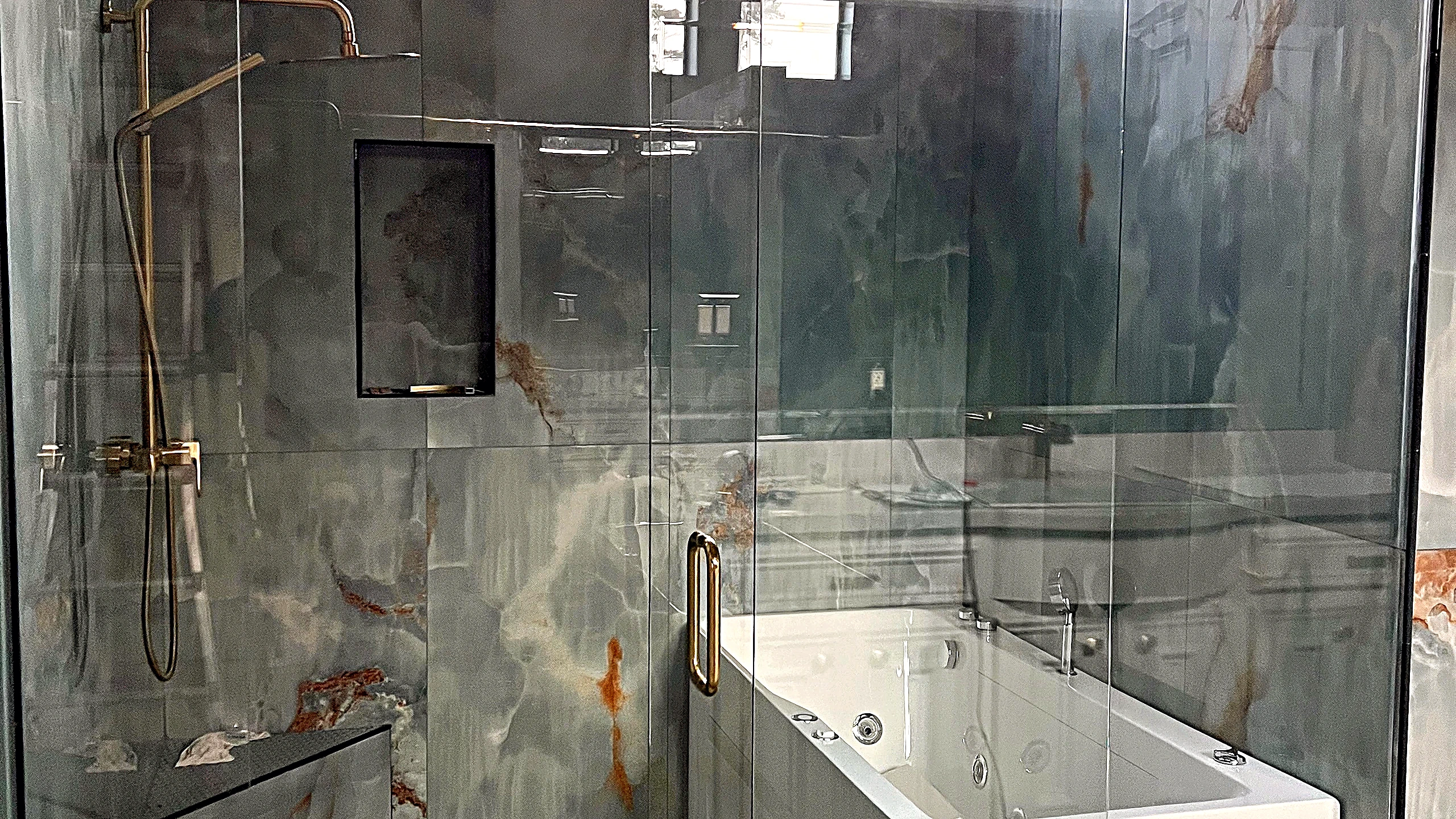 Natural stone slab shower interior with brushed-gold rain shower system, recessed niche, and white soaking tub behind frameless glass