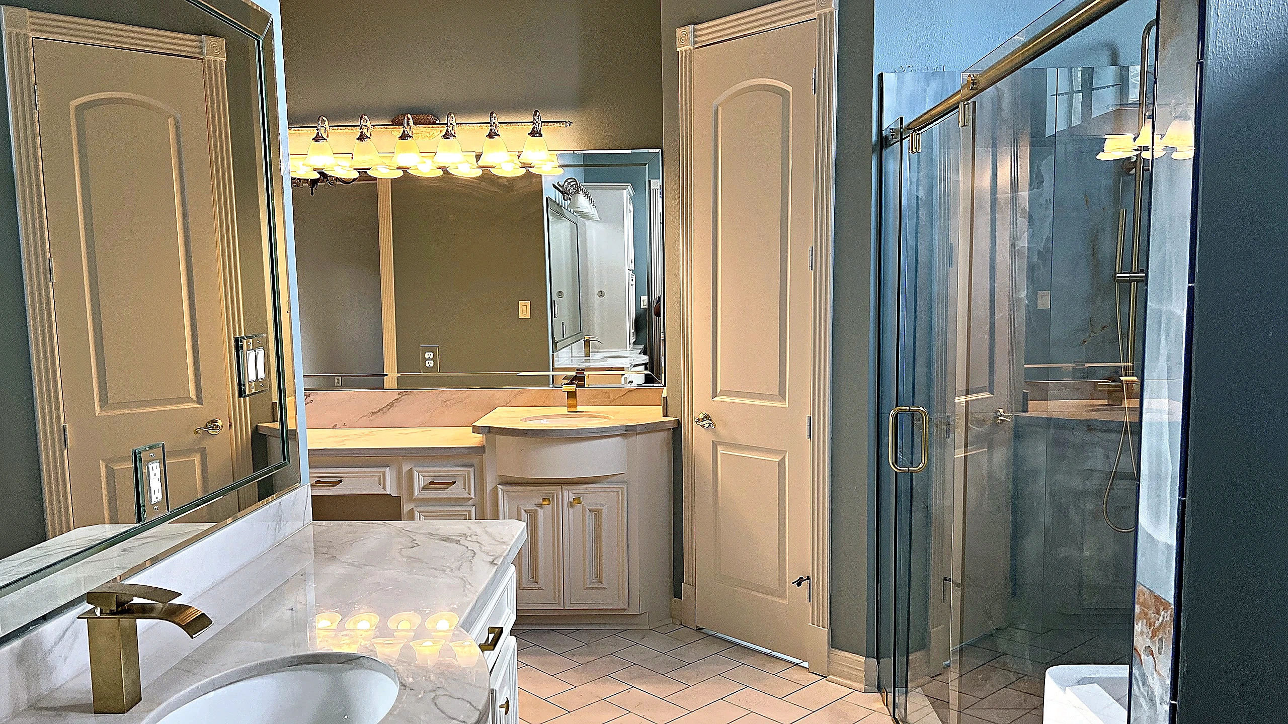 Renovated master bathroom vanity area with white cabinetry, veined stone countertop, gold faucets, and a view into the glass-enclosed shower
