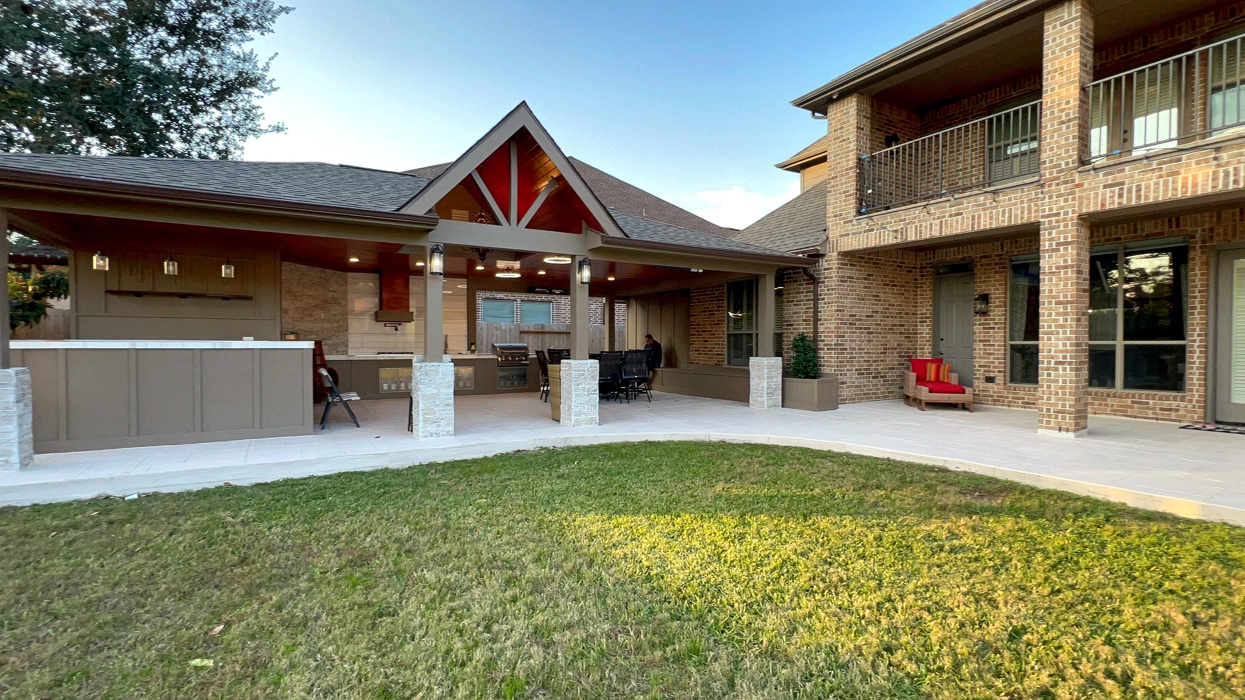 Wide backyard view of completed outdoor kitchen pavilion with pitched roof, stone columns, and bar counter at dusk