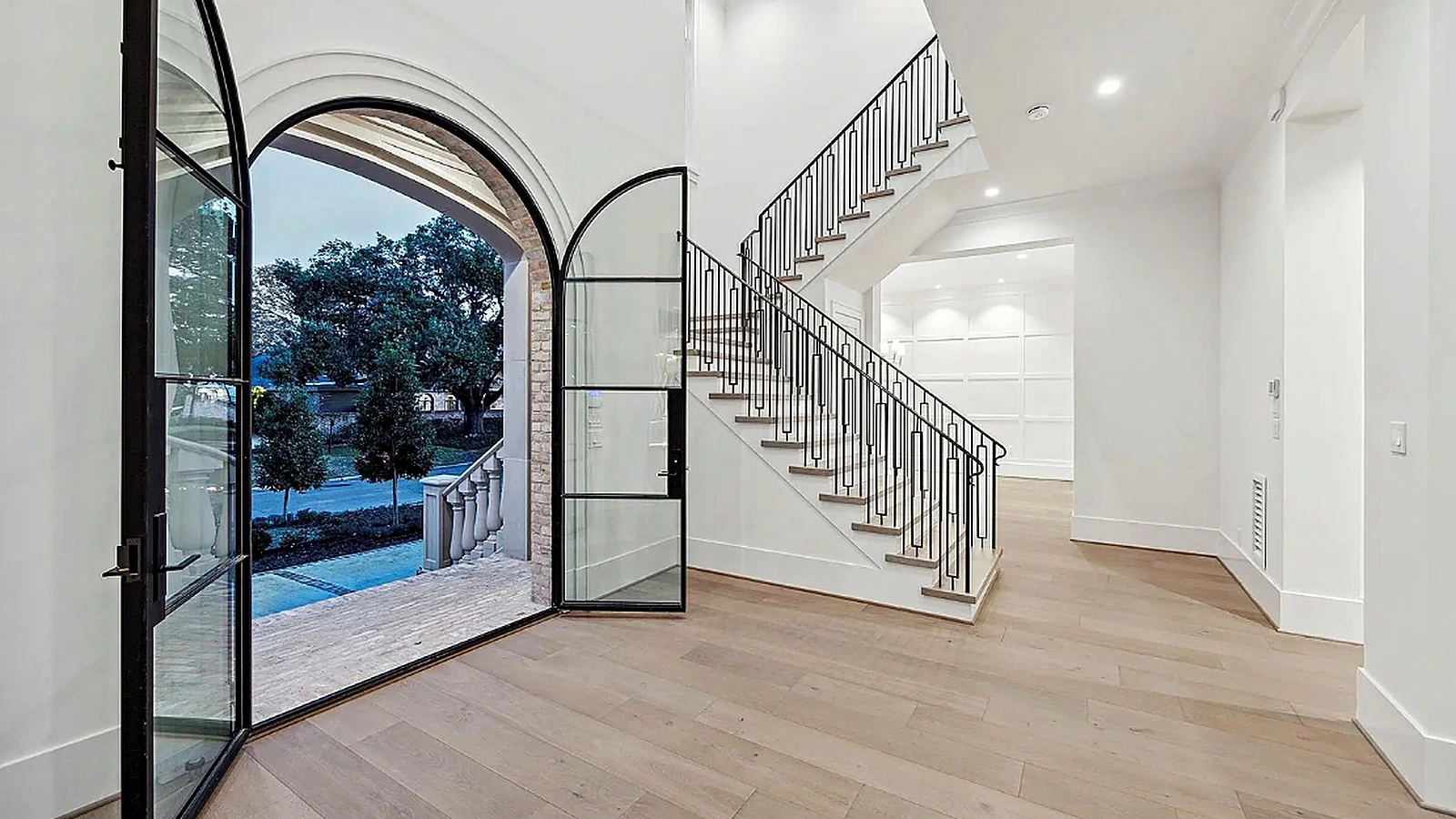 Arched steel-and-glass pivot door open at dusk, stone surround, curved staircase with iron balusters beyond